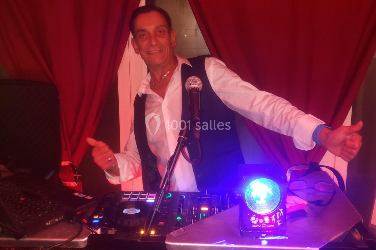 Un DJ souriant derrière une table de mixage, avec un micro et un projecteur lumineux devant des rideaux rouges.