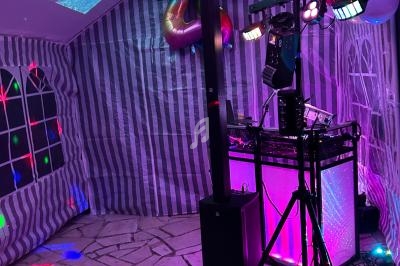 Intérieur d'une tente décorée pour une fête, avec éclairages colorés, équipement DJ et ballons suspendus.