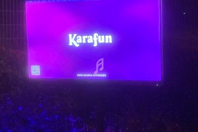 Écran lumineux violet affichant le logo ’Karafun’ et un QR code, placé en extérieur devant une clôture.