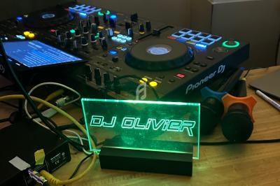 Console de mixage DJ avec éclairage LED vert affichant ’DJ Olivier’, un microphone et des câbles sur une table en bois.