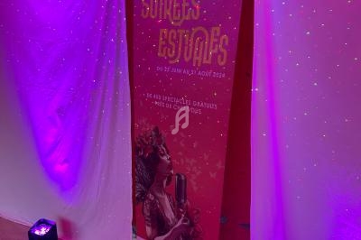 Roll-up coloré annonçant les ’Soirées Estivales’ 2024, entouré de rideaux blancs et éclairages violets.