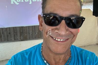 Homme souriant portant des lunettes de soleil et un t-shirt bleu, devant un écran affichant ’Karafun’ en extérieur.