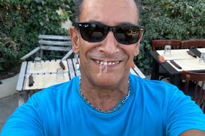 Homme souriant portant des lunettes de soleil et un t-shirt bleu, devant une terrasse de restaurant avec végétation en…