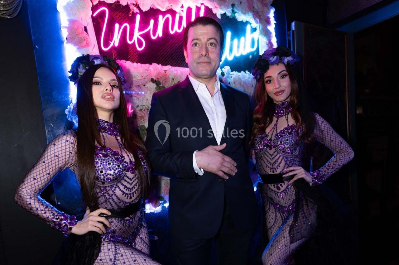 Un homme en costume pose entre deux danseuses en tenues violettes ornées de perles, devant un décor lumineux.