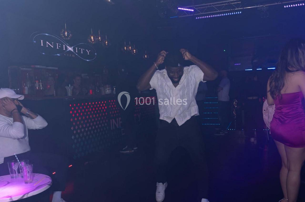 Un homme danse sur une piste de club sombre avec des lumières colorées et un bar en arrière-plan.