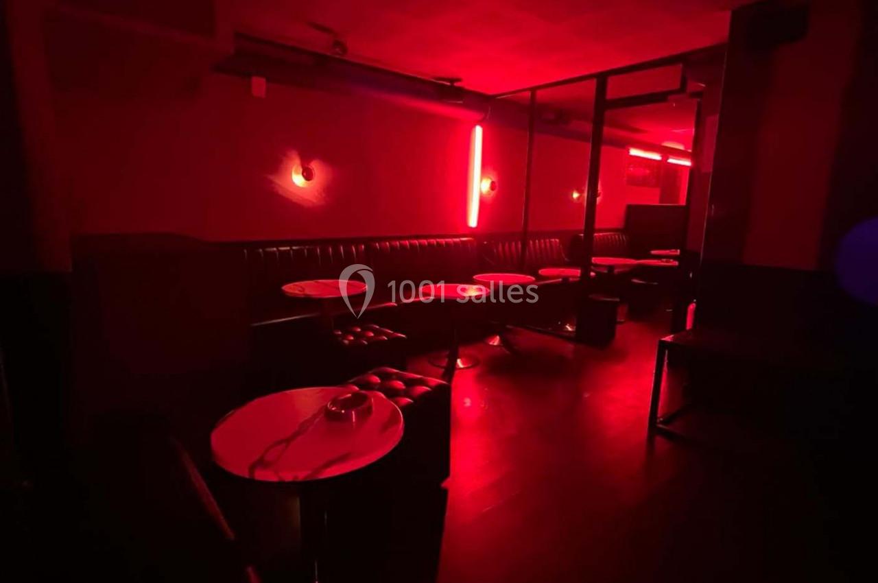 Salle sombre éclairée par des lumières rouges, avec des banquettes, des tables rondes et un sol en bois.