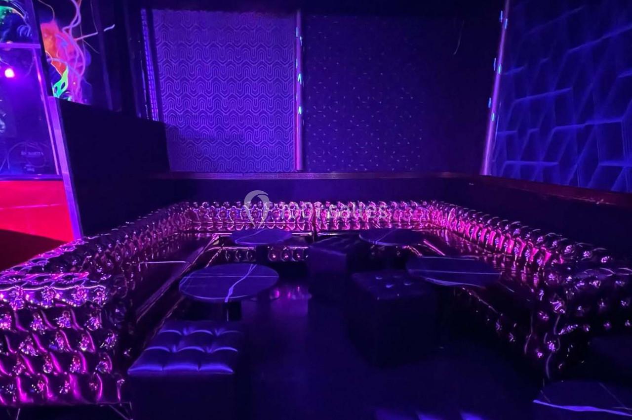 Banquette en cuir capitonné violet entourant des tables basses dans un espace sombre éclairé par des lumières colorées.