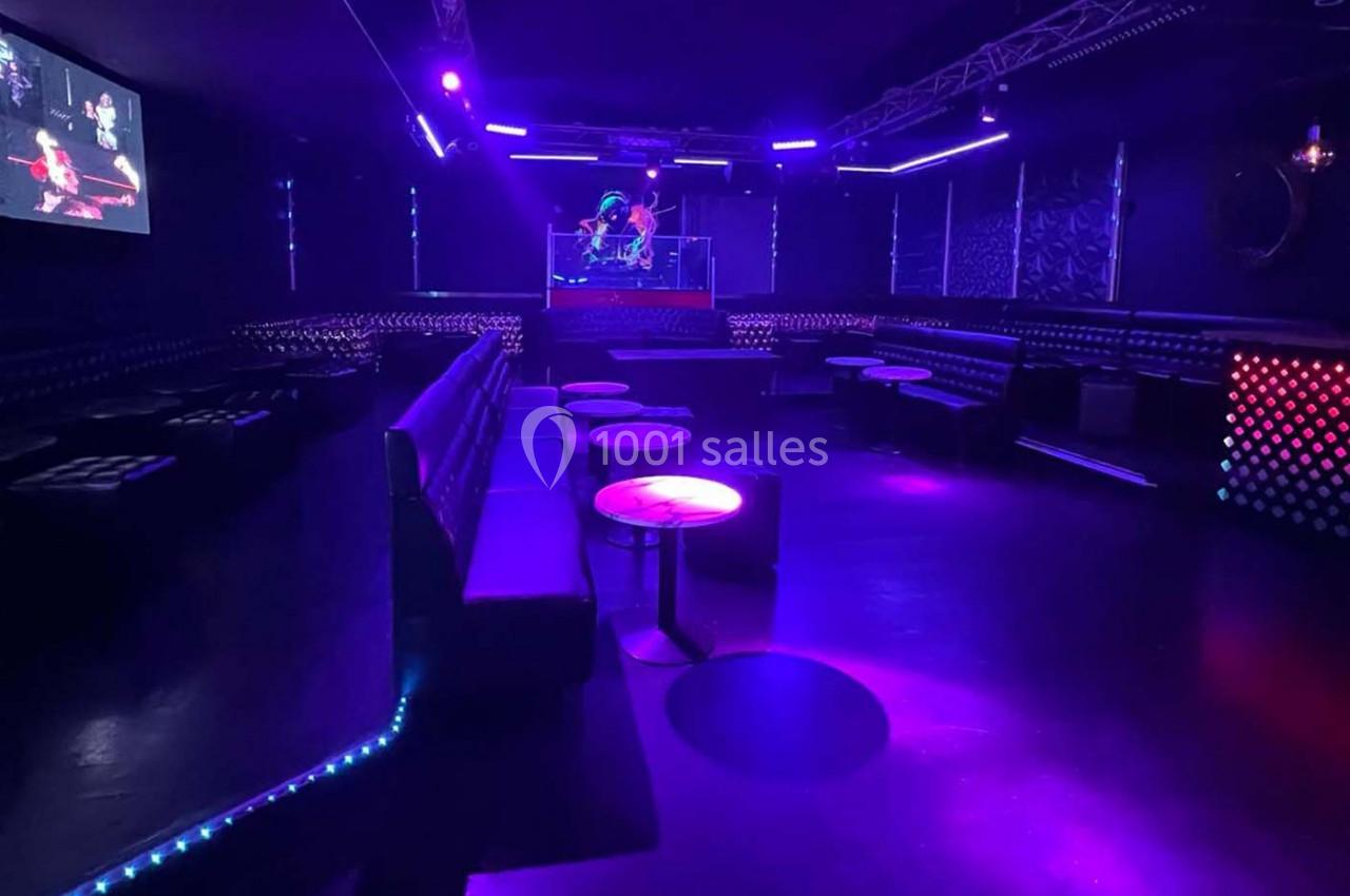 Salle de discothèque vide avec éclairage violet et bleu, banquettes, tables basses et une scène au fond.