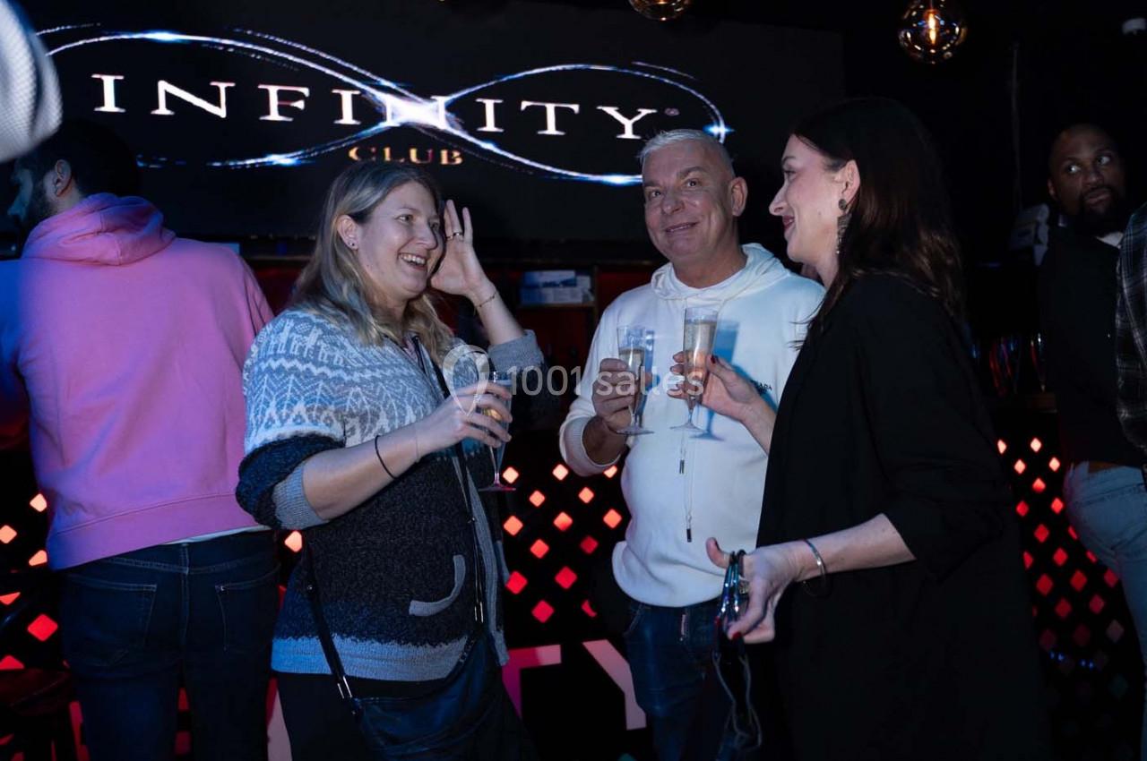 Des personnes discutent et rient en tenant des verres dans un club avec un panneau lumineux ’Infinity Club’ en arrière-plan.