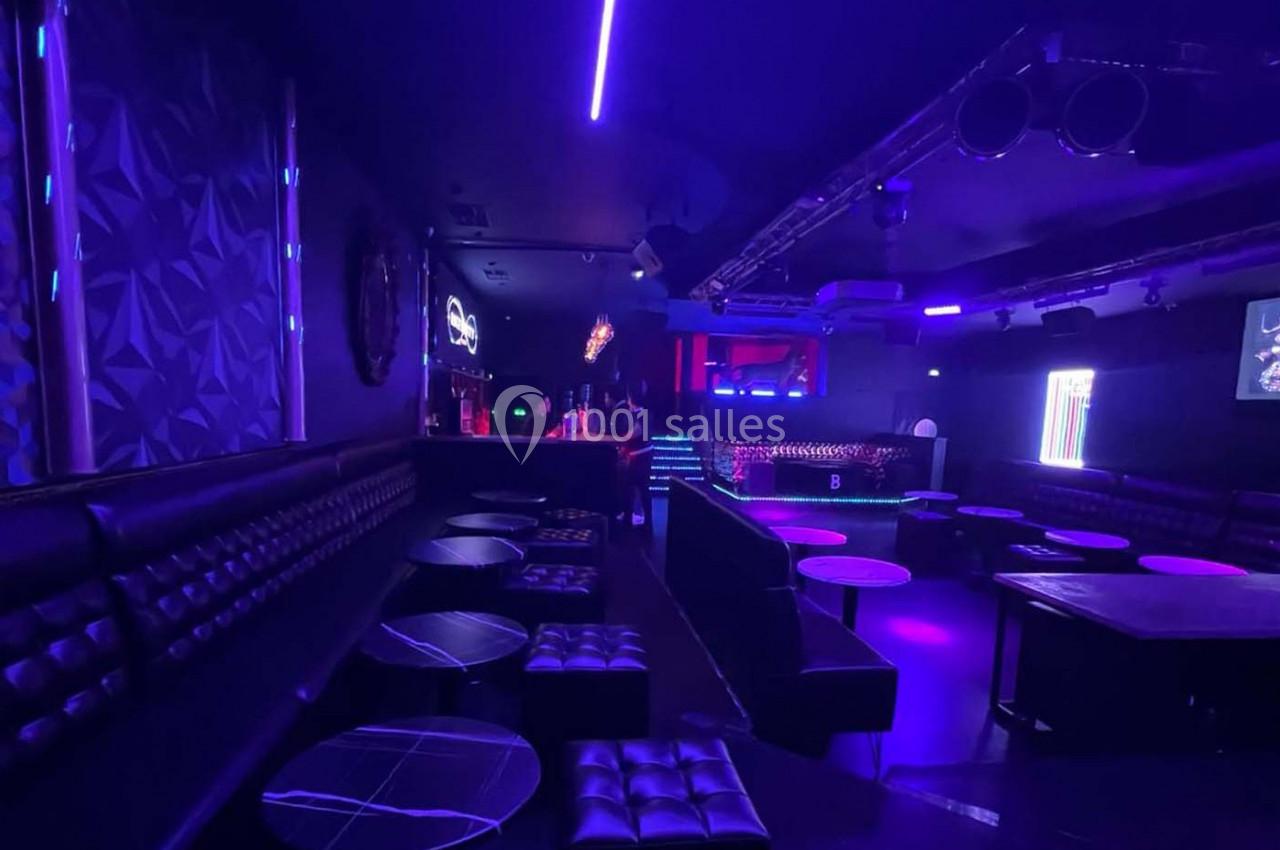 Salle de discothèque sombre avec éclairage violet, banquettes noires, tables basses et piste de danse en arrière-plan.