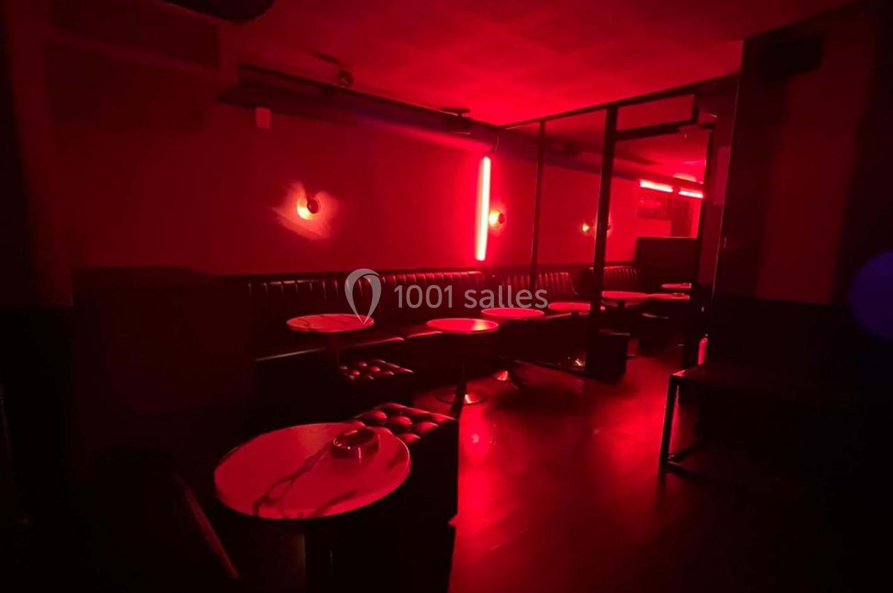 Salle sombre éclairée par une lumière rouge, avec des banquettes en cuir, des tables rondes et un sol en bois.