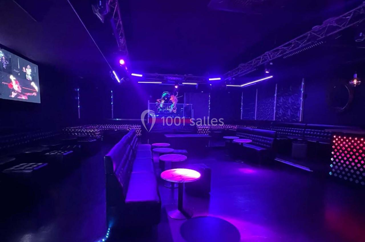 Salle de club vide avec éclairage violet, banquettes noires, tables basses et une scène équipée de lumières.