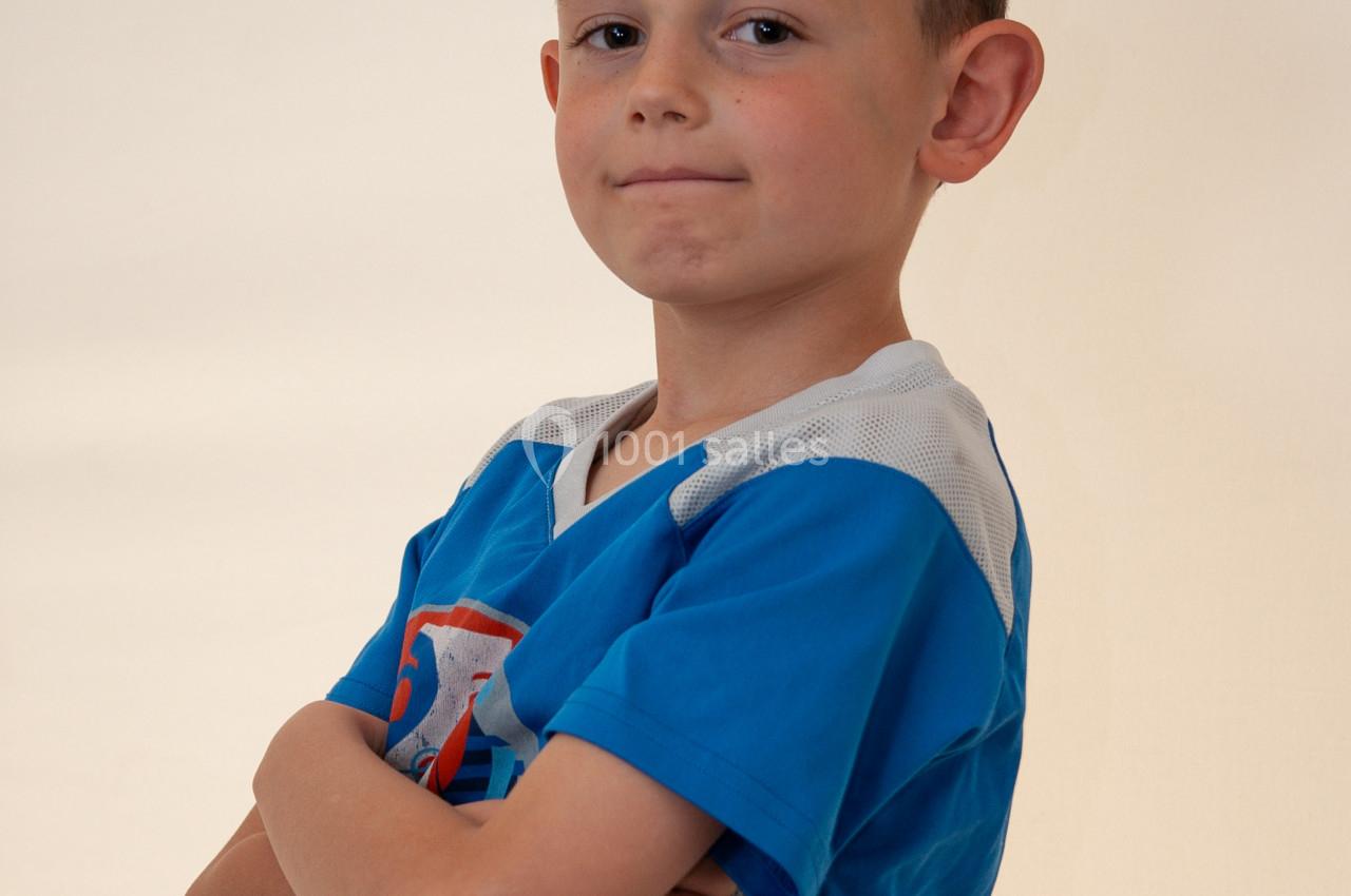Un enfant en t-shirt bleu croise les bras devant un fond clair, regardant l'objectif avec une expression confiante.