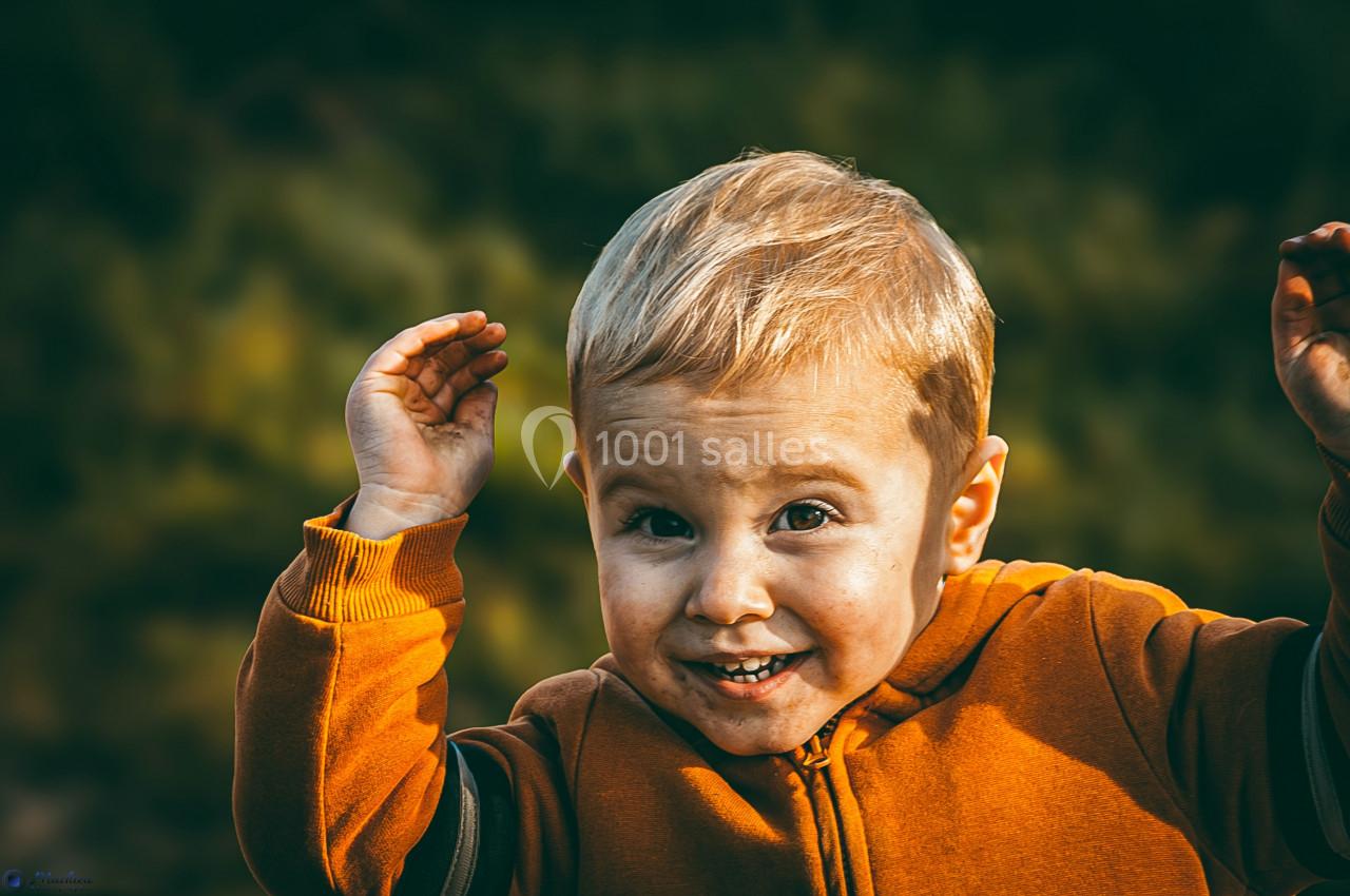 Un enfant blond souriant, vêtu d'un sweat orange, lève les mains devant un fond flou de feuillage.