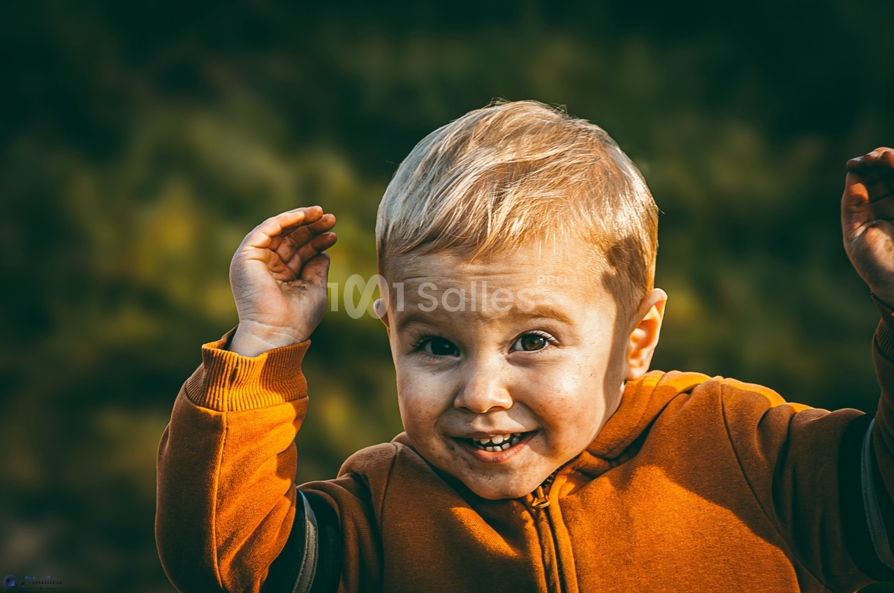 Un enfant blond souriant, vêtu d'un sweat orange, lève les mains devant un fond flou de feuillage.