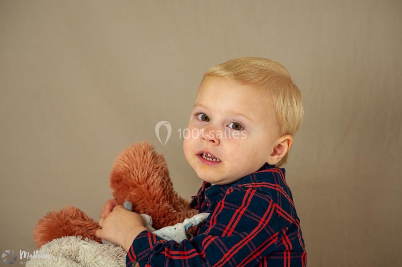 Un jeune enfant blond tenant une peluche marron dans ses bras, sur un fond beige uni.
