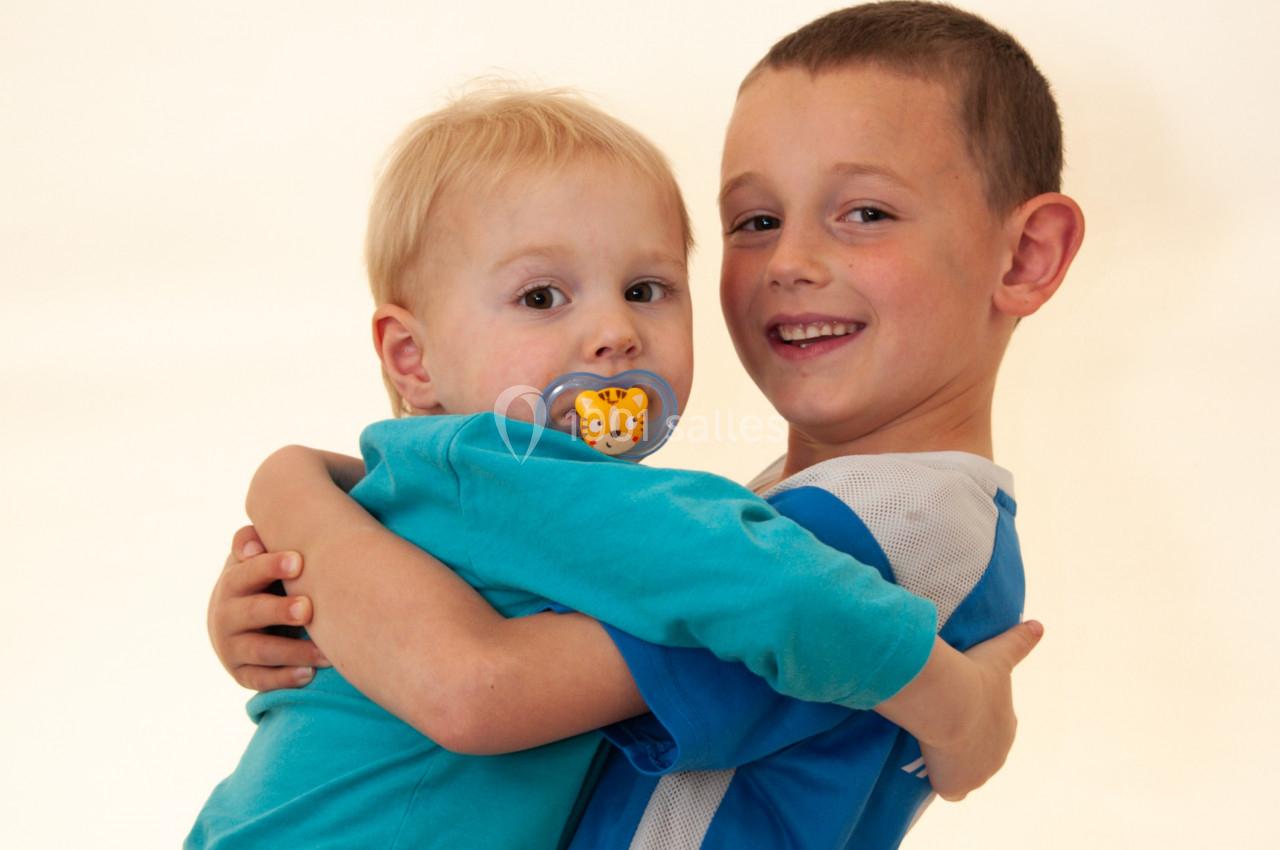 Un enfant plus âgé sourit en portant un jeune enfant avec une tétine, sur un fond clair.