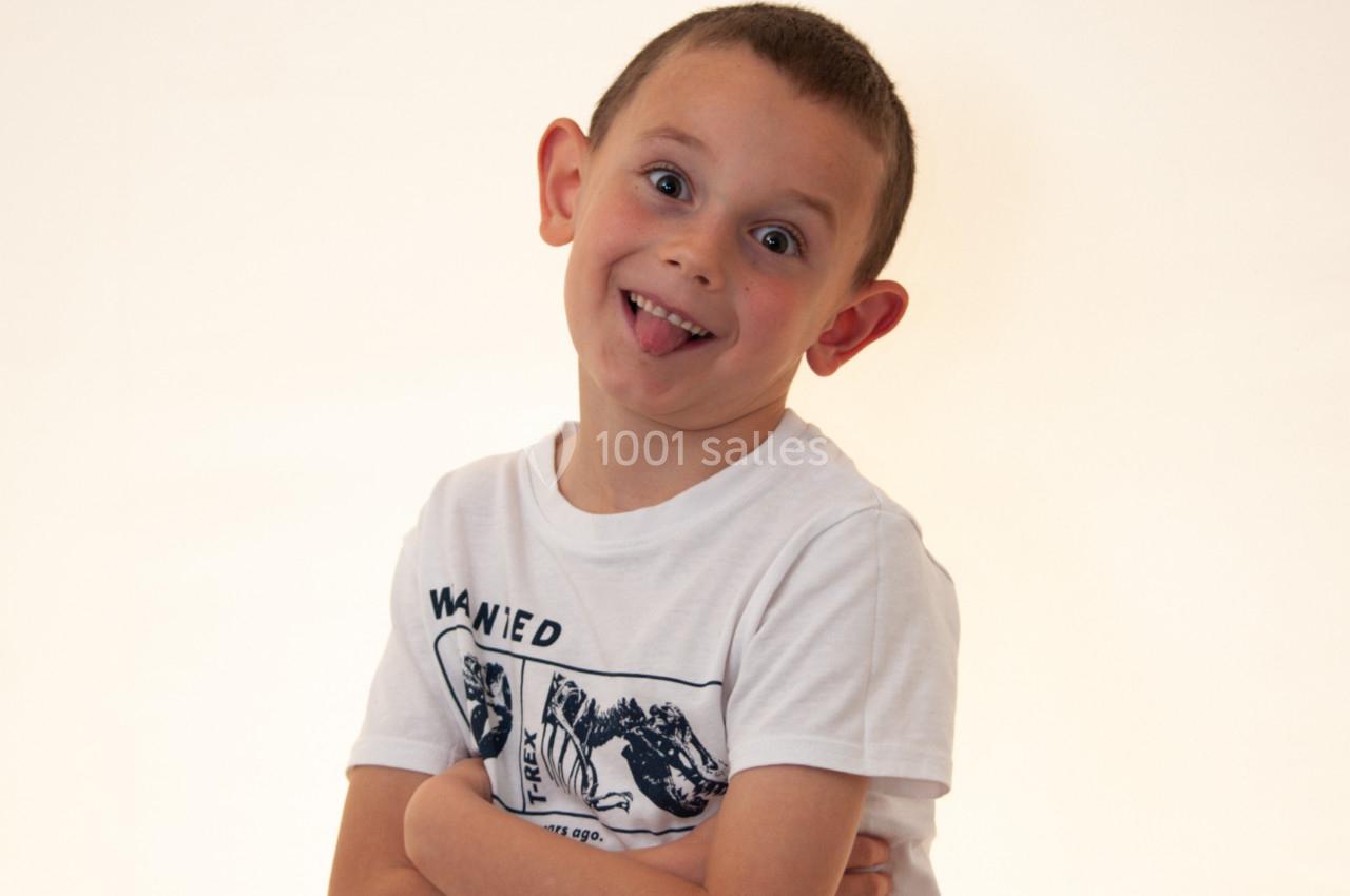 Un enfant souriant, bras croisés, tirant la langue, vêtu d'un t-shirt blanc, sur fond clair.