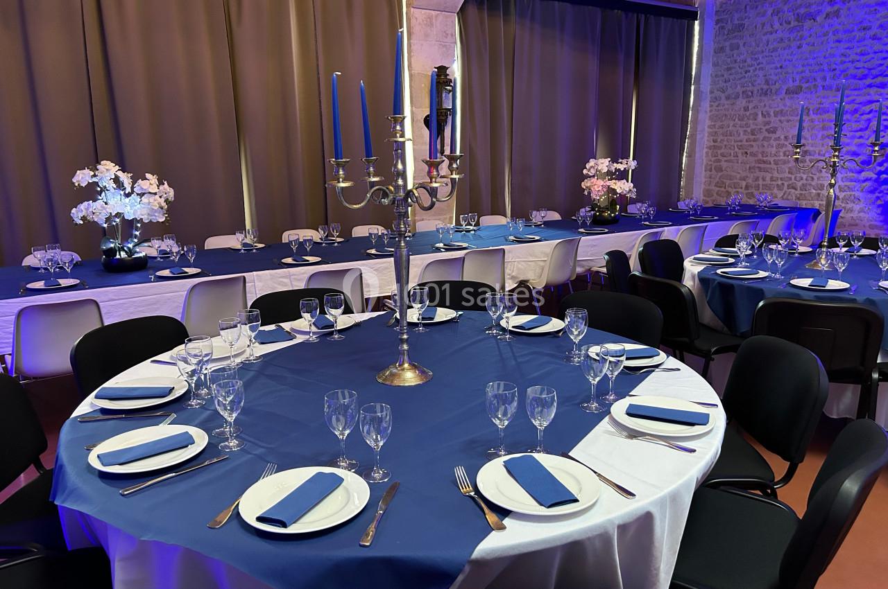 Salle de réception décorée avec des tables rondes dressées, nappes bleues et chandeliers, éclairage tamisé.