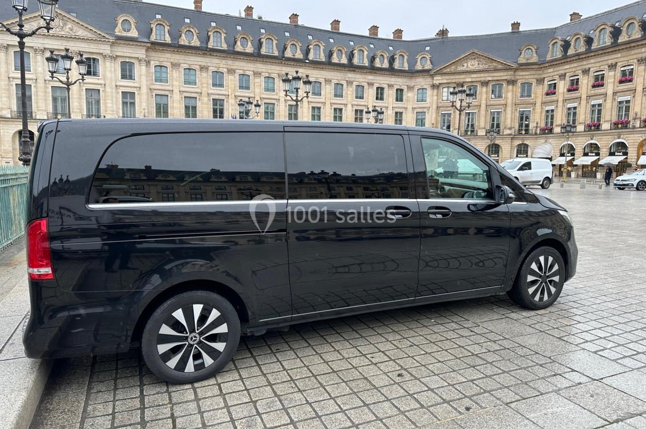 Van noir stationné sur une place pavée devant un bâtiment historique avec des fenêtres alignées et des lampadaires.