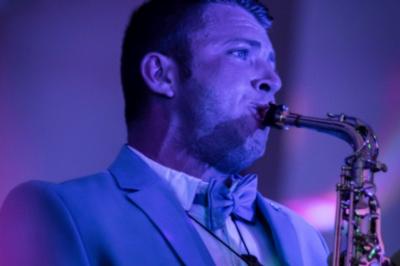 Un homme en costume bleu tient un saxophone sous une pergola décorée de fleurs blanches.