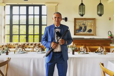 Un homme en costume bleu tient un saxophone sous une pergola décorée de fleurs blanches.