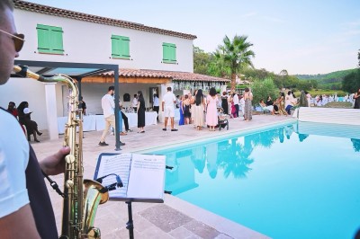 Un homme en costume bleu tient un saxophone sous une pergola décorée de fleurs blanches.