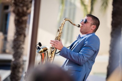 Un homme en costume bleu tient un saxophone sous une pergola décorée de fleurs blanches.
