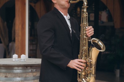 Un homme en costume bleu tient un saxophone sous une pergola décorée de fleurs blanches.