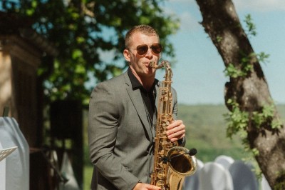 Un homme en costume bleu tient un saxophone sous une pergola décorée de fleurs blanches.