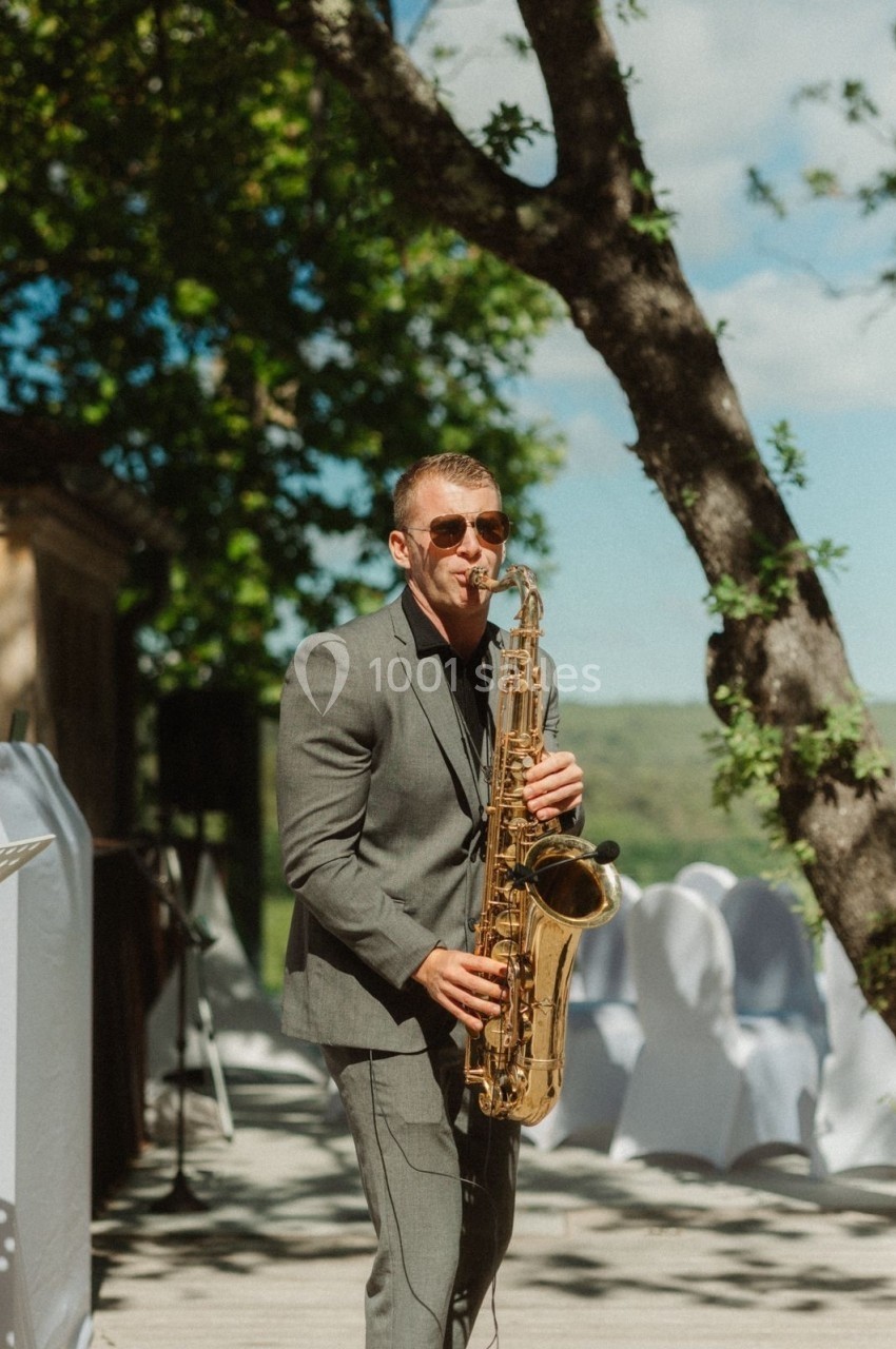 Un musicien en costume joue du saxophone en extérieur, sous un arbre, par une journée ensoleillée.