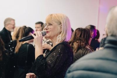 Femme blonde tenant un micro, parlant devant un groupe de personnes dans un espace intérieur éclairé.