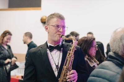 Un musicien en smoking joue du saxophone lors d'un événement avec des personnes en arrière-plan.