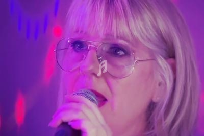 Femme blonde portant des lunettes, chantant au micro sous des lumières violettes et rouges.