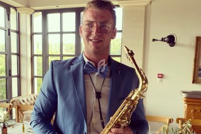 Un homme en costume bleu tient un saxophone dans un intérieur lumineux avec de grandes fenêtres.
