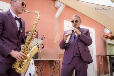 Deux musiciens en costume violet jouent du saxophone et d'un instrument à vent devant un bâtiment orange.