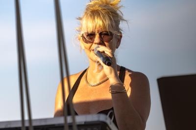 Femme blonde portant des lunettes de soleil chantant au micro devant un ciel dégagé avec un clavier en avant-plan.