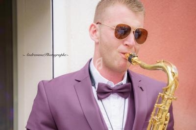 Un homme en costume violet joue du saxophone devant un mur beige et rose.