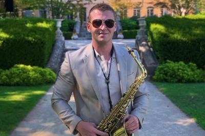 Un homme en costume beige tenant un saxophone dans un jardin avec des haies et un bâtiment en arrière-plan.