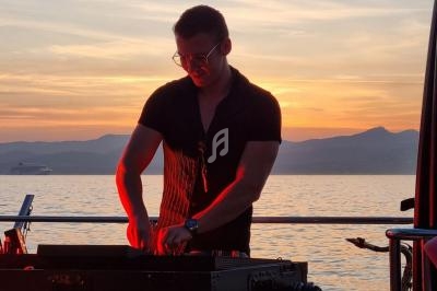 Un DJ mixe sur un bateau au coucher du soleil, avec une vue sur la mer et des montagnes en arrière-plan.