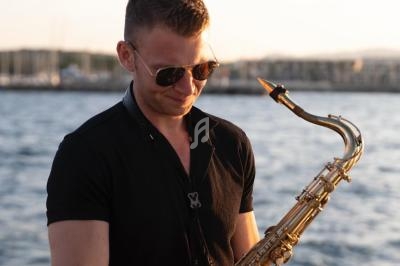 Un homme en chemise noire joue du saxophone devant un plan d'eau au coucher du soleil.