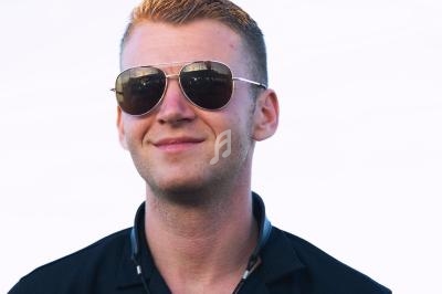 Homme souriant portant des lunettes de soleil et une chemise noire, sur un fond blanc uni.