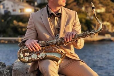 Un homme en costume beige joue du saxophone assis sur des rochers près d'une étendue d'eau au coucher du soleil.