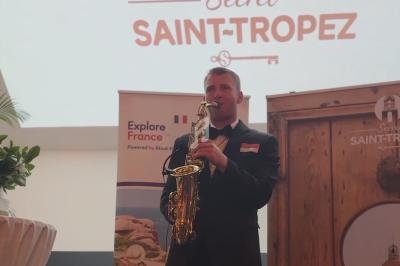 Un musicien joue du saxophone devant des affiches promotionnelles de Saint-Tropez dans un espace intérieur.