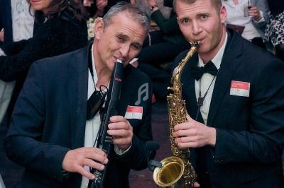 Deux musiciens en costume jouent du saxophone et d'un instrument à vent électronique lors d'un événement en intérieur.