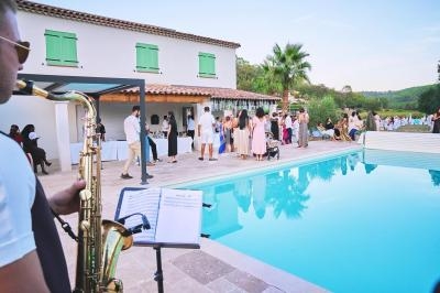 Un saxophoniste joue près d'une piscine lors d'un événement en plein air avec des invités rassemblés autour.