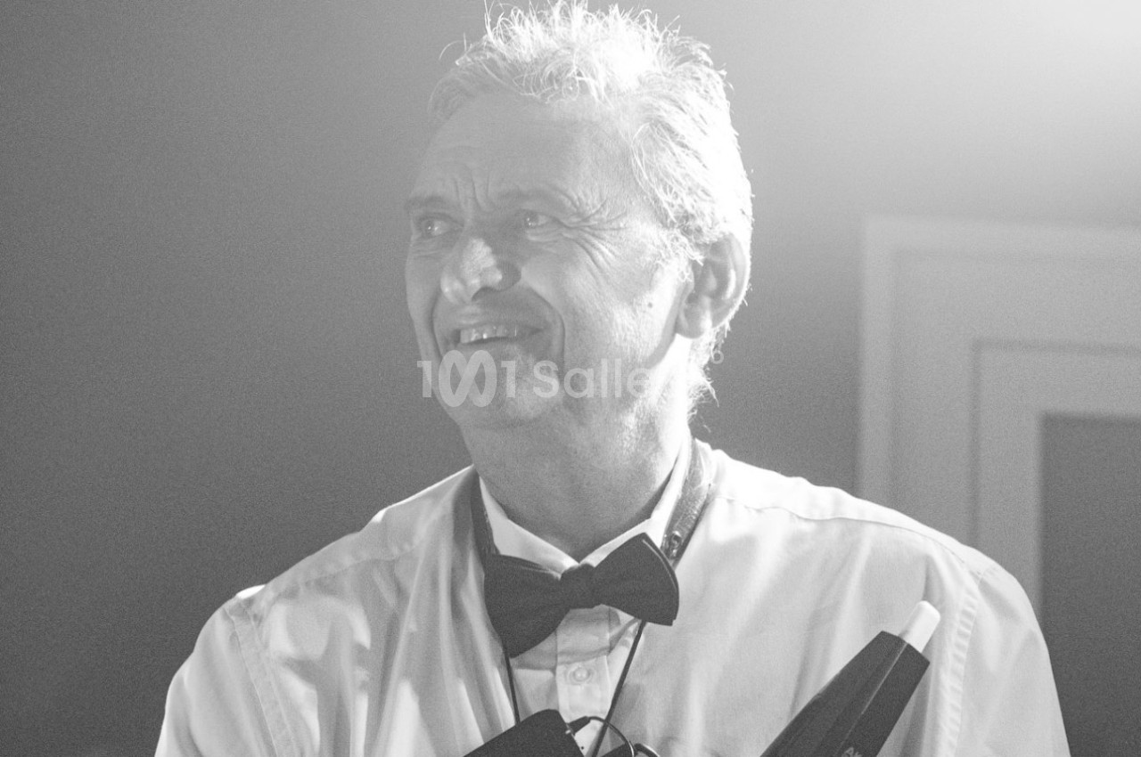 Homme souriant en chemise blanche et nœud papillon, éclairé par une lumière diffuse en noir et blanc.