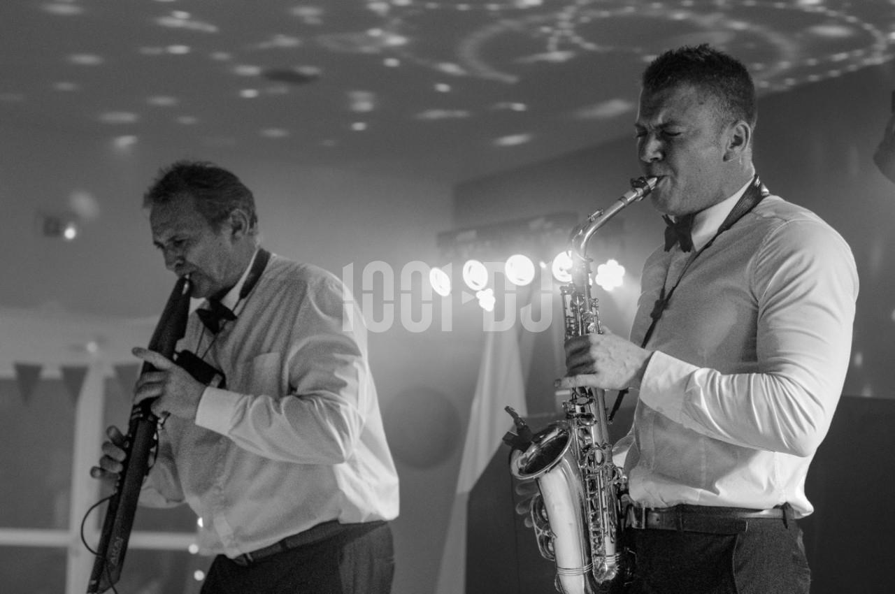 Deux musiciens en chemises blanches jouent du saxophone et d'un instrument à vent dans une ambiance éclairée de spots.