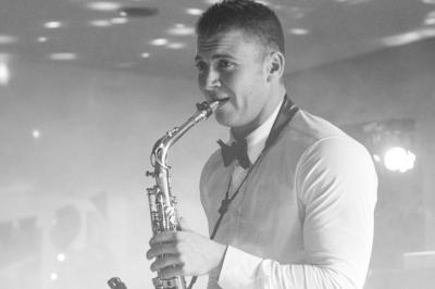 Un homme en costume bleu tient un saxophone dans un intérieur lumineux avec de grandes fenêtres.