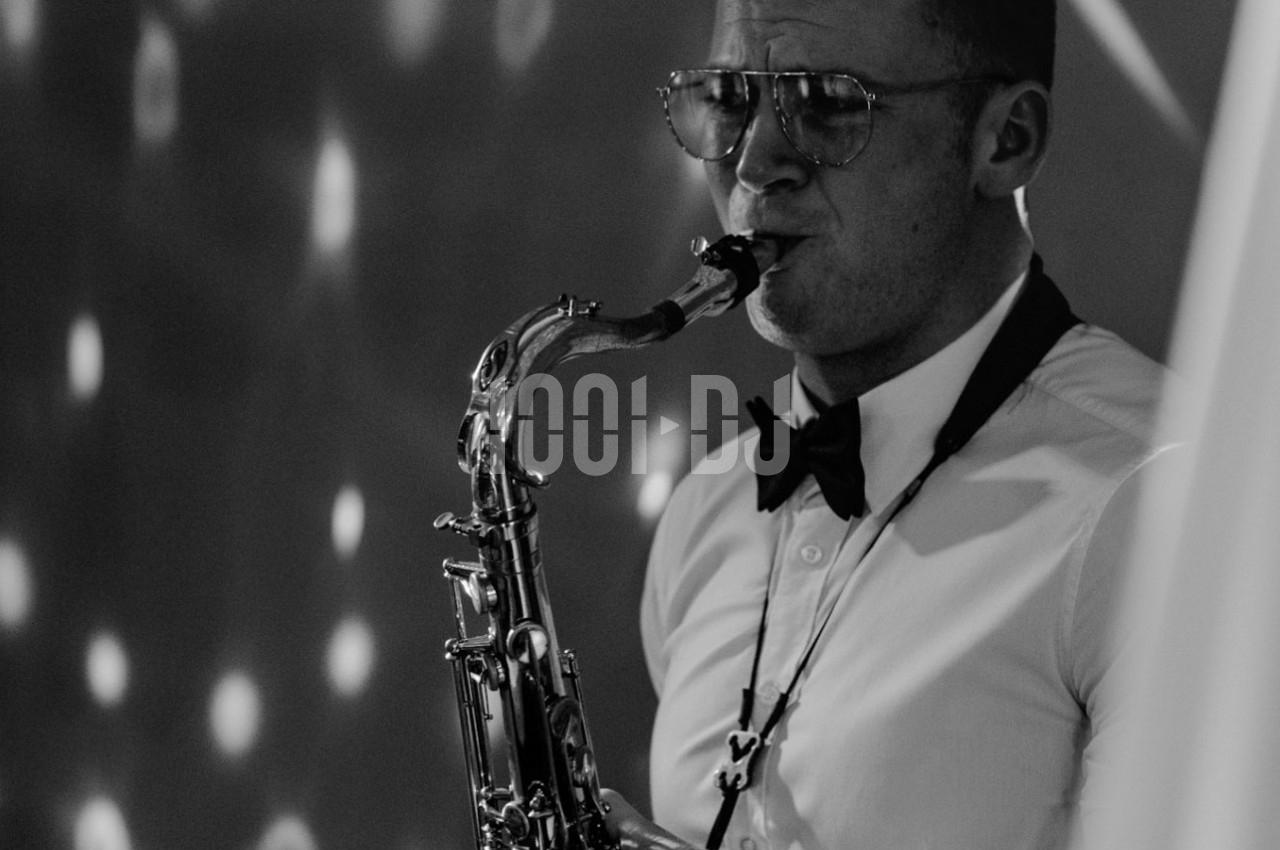 Un musicien en chemise et nœud papillon joue du saxophone dans une ambiance éclairée de lumières tamisées.
